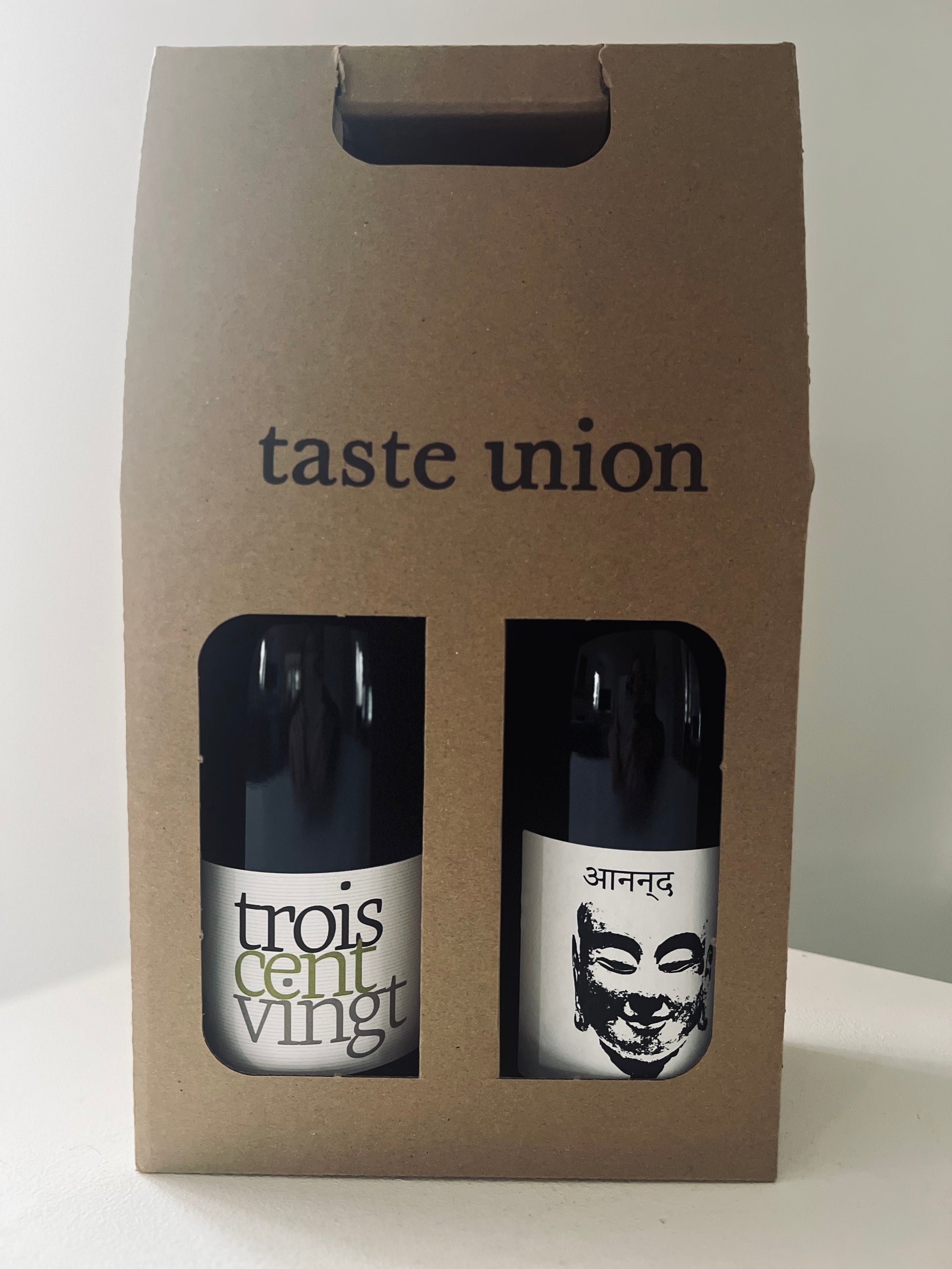 A cardboard gift box from Taste Union containing two bottles of natural wine, visible through cut-out windows. The left bottle is Domaine Pion Trois Cent Vingt, featuring a minimalist white label with bold typography. The right bottle is Mouton Noir Ananda, showcasing a white label with a black Buddha face design and the word 'Ananda' in a script font.