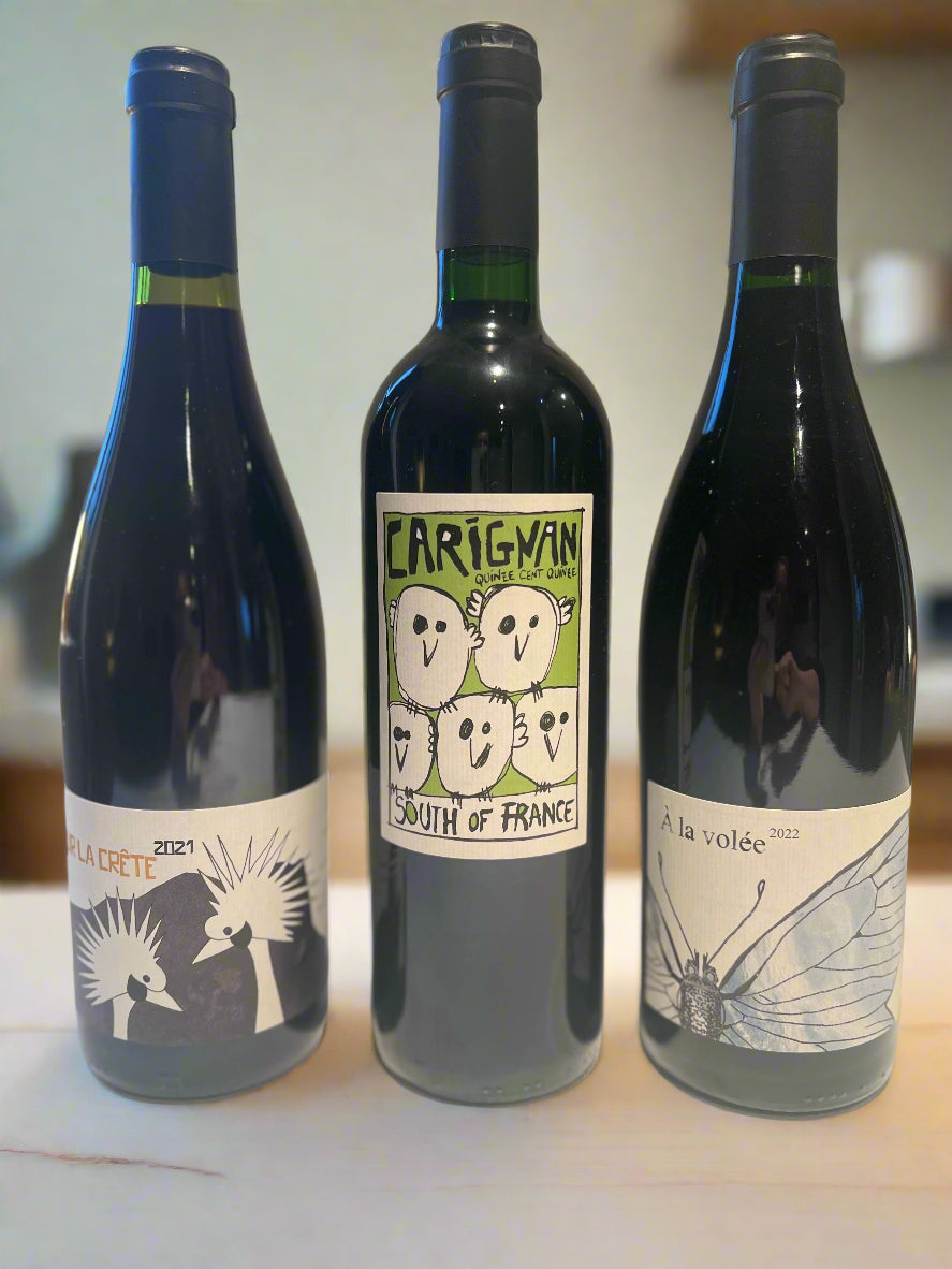 Three bottles of natural red wine from Colline de l’Hirondelle: La Crête 2021, Carignan South of France, and À la volée 2022, displayed on a light wooden surface.