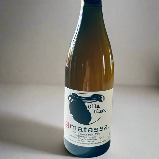 A bottle of Matassa Olla Blanc 2021, a biodynamic natural white wine made with Muscat Petit Grain and Macabeu grapes, standing on a white surface with a grey background.