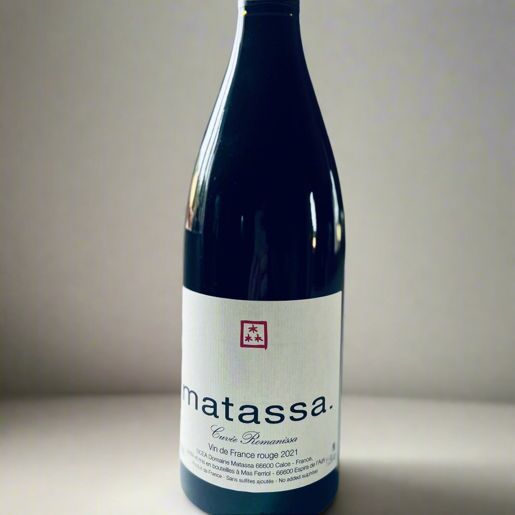 A bottle of Matassa Romanissa Organic Red Wine 2021, featuring a minimalist white label with black text and a red logo. Positioned on a white surface with soft, natural lighting in the background.
