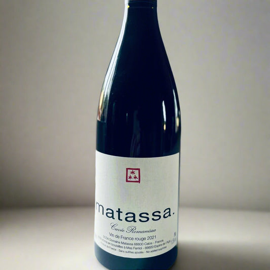 A bottle of Matassa Romanissa Organic Red Wine 2021, featuring a minimalist white label with black text and a red logo. Positioned on a white surface with soft, natural lighting in the background.